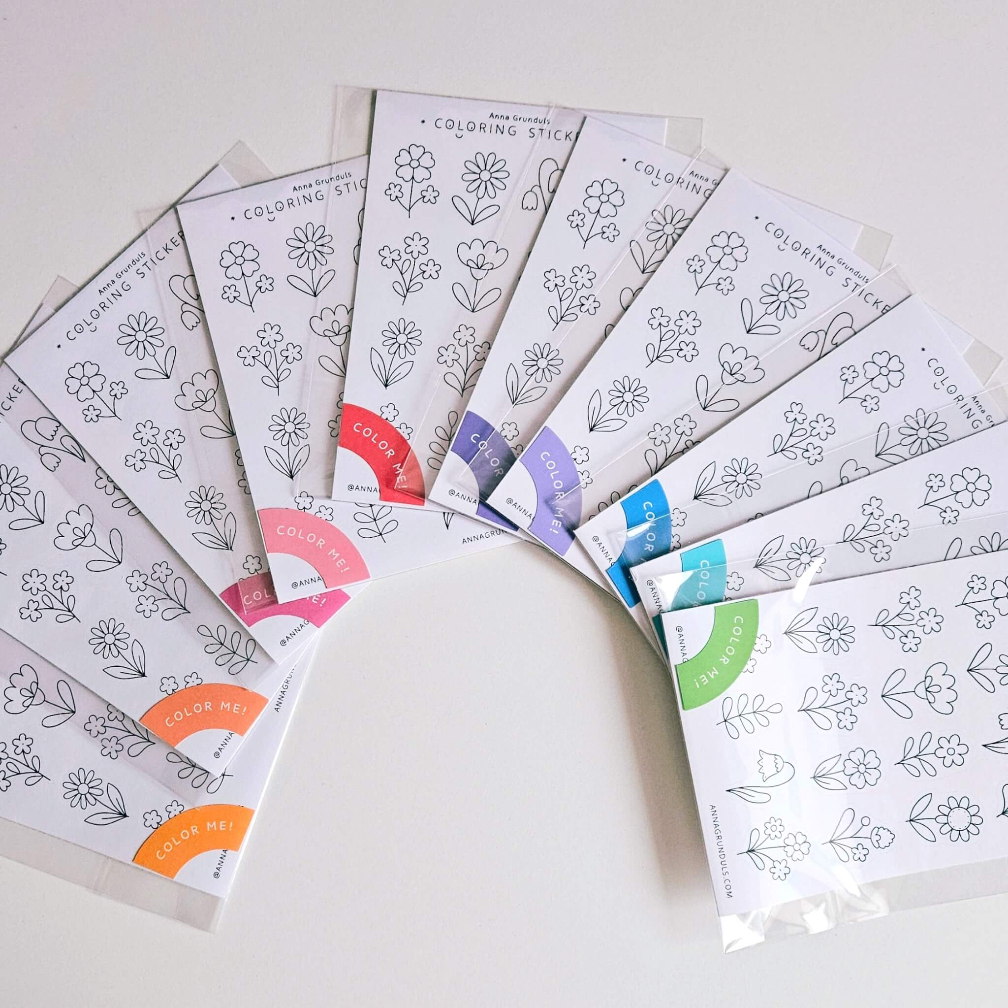Several floral coloring sticker sheets arranged in a fan shape, each featuring hand-drawn black and white flower illustrations. The A6 sticker sheets are packaged in clear sleeves with colorful “Color Me!” labels and are designed to be colored in like a mini coloring page.