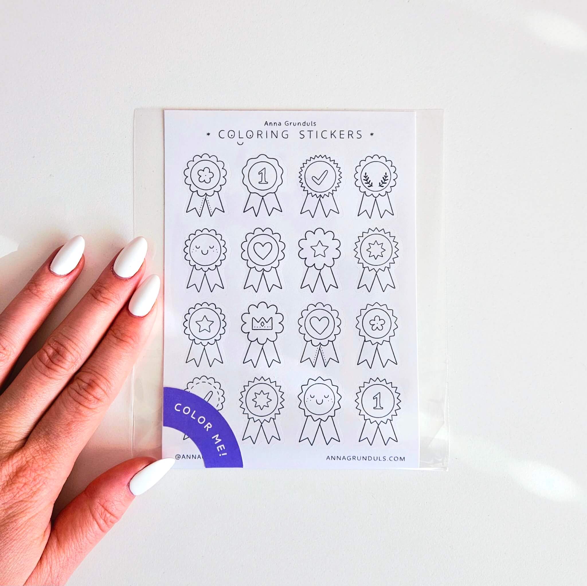 An award ribbon coloring sticker sheet featuring hand-drawn black and white rosette badge designs with stars, hearts, crowns, check marks, and number one symbols. A hand with light neutral manicure holds the A6 sticker sheet, which is packaged in a clear sleeve with a purple “Color Me!” label in the corner.