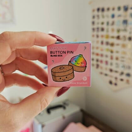 A hand holds a pink viral squishy dumpling button pin blind box decorated with a rainbow dumpling and steamer basket.
